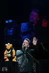 Ventriloquist onstage performing with a turtle puppet in front of a large screen projection.