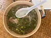 Overhead view of a bowl of pho with herbs and a plastic spoon on a wooden table.