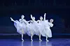 Four ballet dancers in white tutus strike high leg poses onstage in a comic formation.