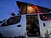 White camper van with open rooftop tent and red string lights at dusk.