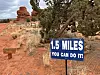 Blue motivational sign beside a desert trail with red rock formations and trees in the background.