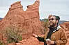 A man in sunglasses stands in front of red sandstone cliffs and gestures while holding a coffee cup.
