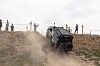 An off-road vehicle drives up a dusty hillside while people stand behind a rope barrier.