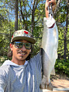 A man in sunglasses and a cap holds up a large trout outdoors near trees and brush.