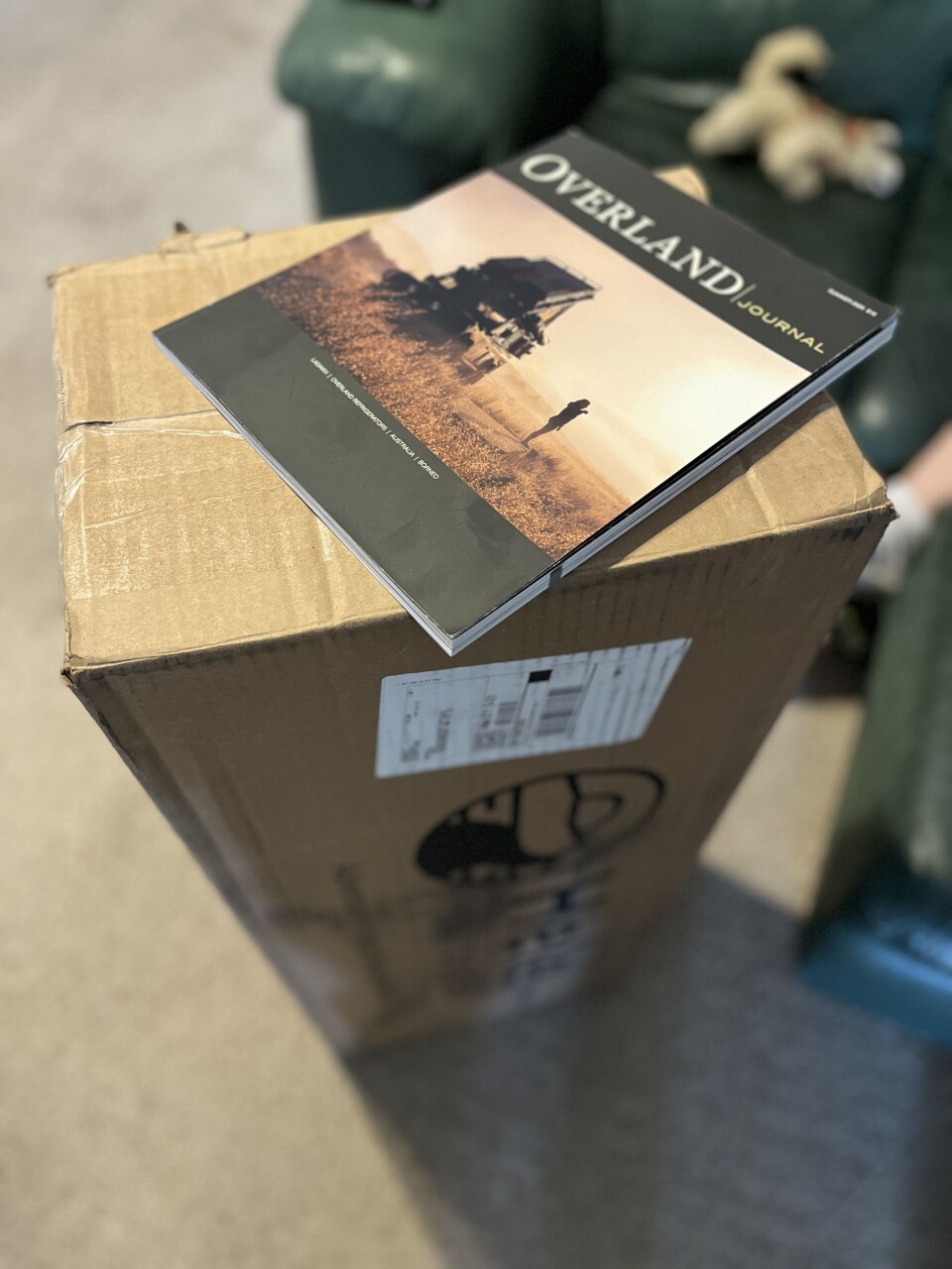 A cardboard shipping box with an Overland journal magazine placed on top in a room.