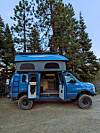 Blue camper van with pop-top roof, open doors and rugged tires parked near pine trees.