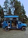 Blue camper van with pop-top roof, open doors and rugged tires parked near pine trees.