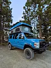 Blue lifted van with a pop-up roof, roof rack, and off-road tires parked in a forest clearing.