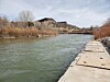 A river flows past a concrete diversion platform with brushy banks and a rocky hill under a cloudy sky.