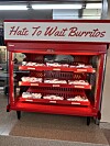 Red refrigerated display case holding packaged breakfast burritos under a sign that reads, 'Hate To Wait Burritos.'