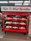 Red refrigerated display case holding packaged breakfast burritos under a sign that reads, 'Hate To Wait Burritos.'