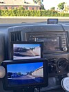 Two screens on a vehicle dashboard show a rear-view camera image of a road behind the vehicle.