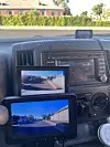 Two screens on a vehicle dashboard show a rear-view camera image of a road behind the vehicle.