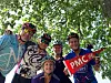 Five cyclists wearing helmets pose outdoors and hold a red Pan-Mass Challenge flag under green tree branches.