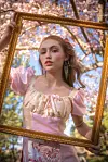 Cassidy Kurowski in a pink dress holding a gold ornate frame outdoors with flowering trees behind her.