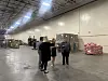 Three people stand in a large warehouse beside stacked pallets of food bags and supplies.