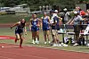 A runner in blue sprints on a red track while several people watch nearby.