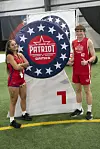 Victoria Perea and Keller Ford stand in red uniforms on indoor turf beside a large Patriot Games banner with a 7.