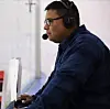 A broadcaster wearing a headset works at a rinkside control station beside the ice.